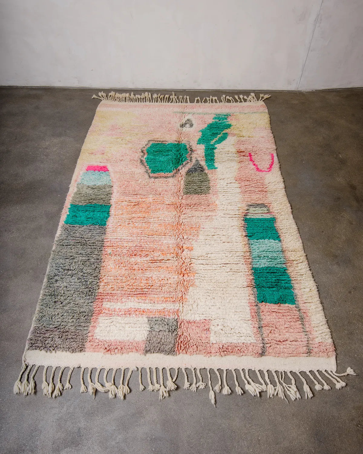 Handcrafted Berber rug from Morocco laying flat on concrete floor. Vintage carpet featuring shades of pink, peach, yellow and turquoise and a fluffy texture.